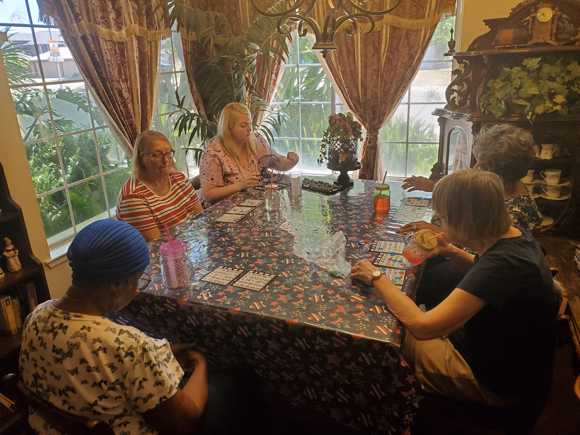 Five elderly women sitting around a table playing bingo in a well-lit room with large windows and decorative curtains. The table is covered with a festive tablecloth and has bingo cards, markers, and drinks on it. There are plants and a wooden cabinet with teacups in the background.