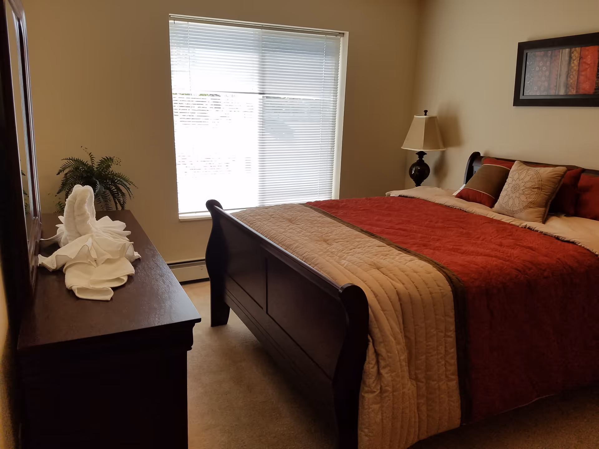 A bedroom with a large wooden bed featuring a red and beige quilt and multiple pillows. There is a wooden dresser with a towel folded into a swan shape and a potted plant on top. A window with blinds lets in natural light, and a lamp sits on a bedside table next to the bed. A framed piece of artwork hangs on the wall above the bed.