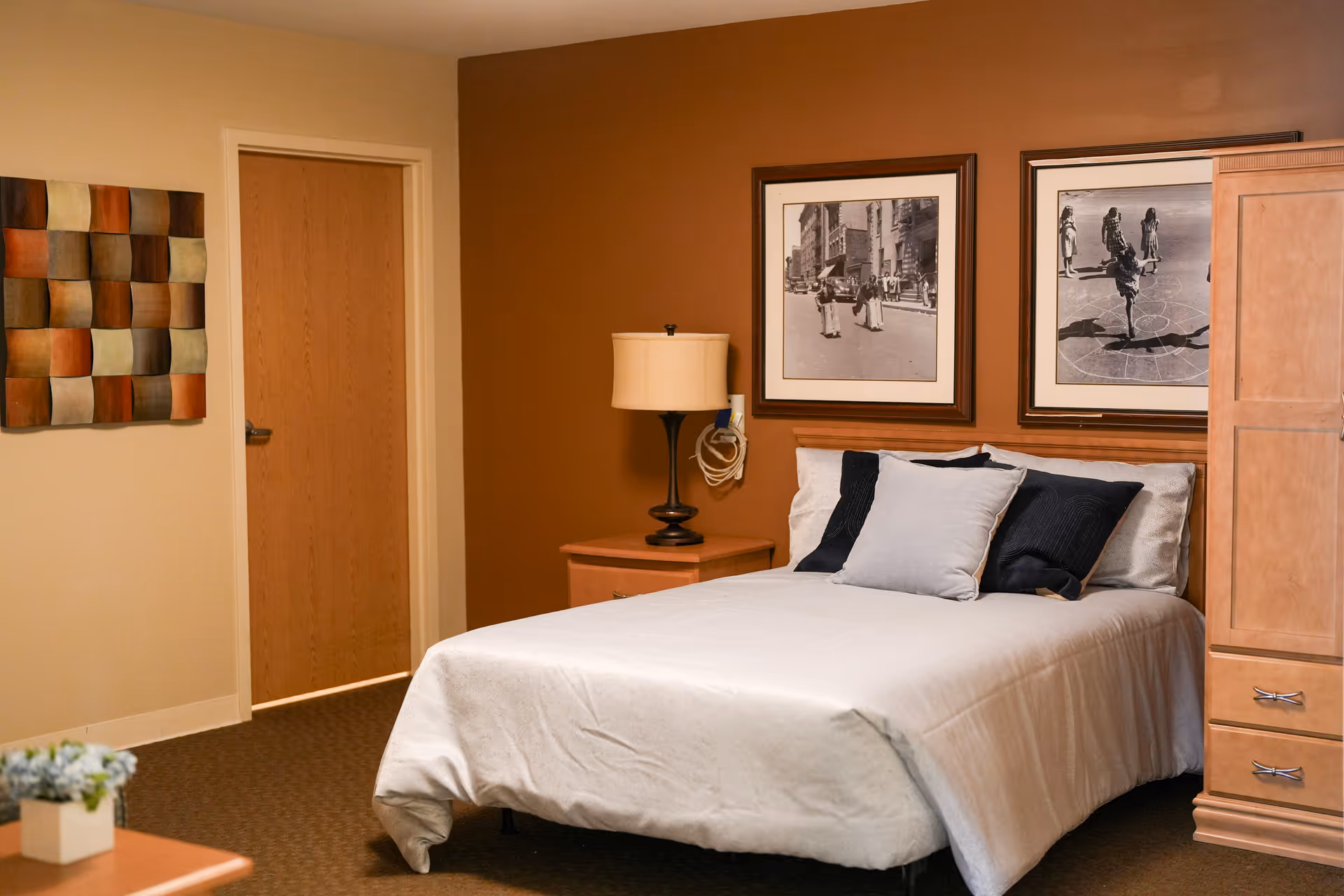 A cozy bedroom with a neatly made bed featuring white and dark blue pillows. The room has warm brown and beige walls, a wooden nightstand with a lamp, two framed black and white photographs above the bed, a wooden wardrobe, and a decorative wall piece near a closed wooden door.