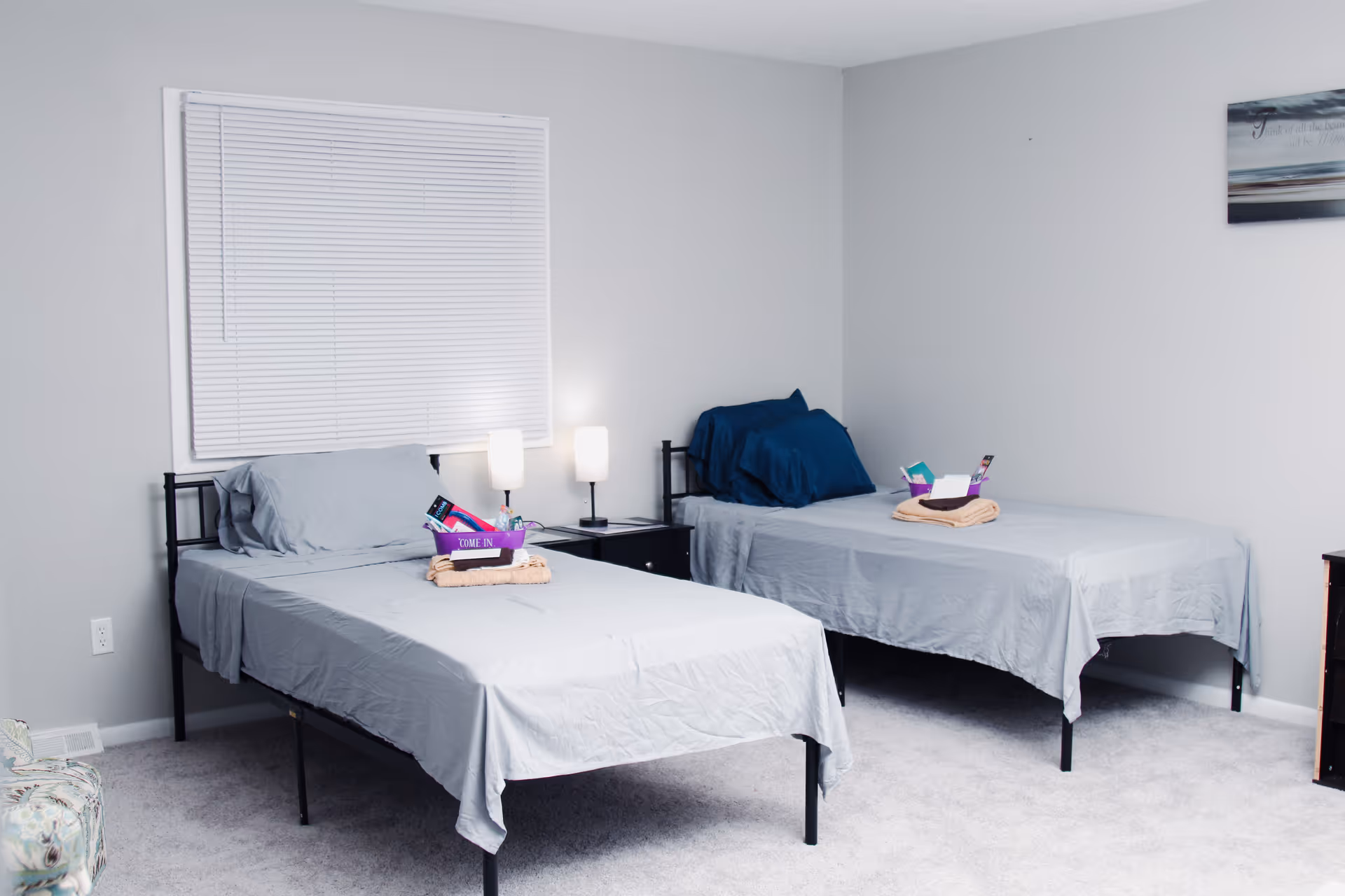 A simple bedroom with two single beds, each made with light gray sheets and a pillow. Each bed has a folded towel and a small basket with toiletries placed on top. There is a nightstand with two lamps between the beds, a window with closed blinds behind one bed, and a piece of wall art on the right wall. The room has light gray walls and carpeted floor.