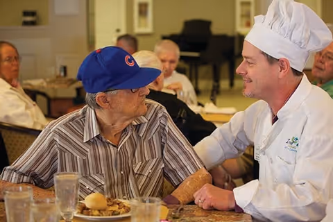 An elderly man wearing a blue baseball cap and striped shirt sits at a dining table with a plate of food in front of him. He is looking and smiling at a chef dressed in white with a chef's hat, who is sitting beside him and engaging in conversation. Other elderly people are seated in the background in a communal dining area.