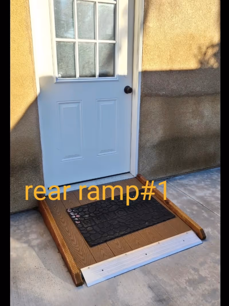 A small wooden ramp leading up to a white door with glass panes, placed against a beige stucco wall. A black welcome mat is positioned on the ramp. The text 'rear ramp#1' is overlaid on the image in yellow.