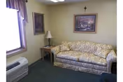 Small sitting room with a floral couch, side table and lamp, framed pictures and a crucifix on the wall, and a window with an air unit.