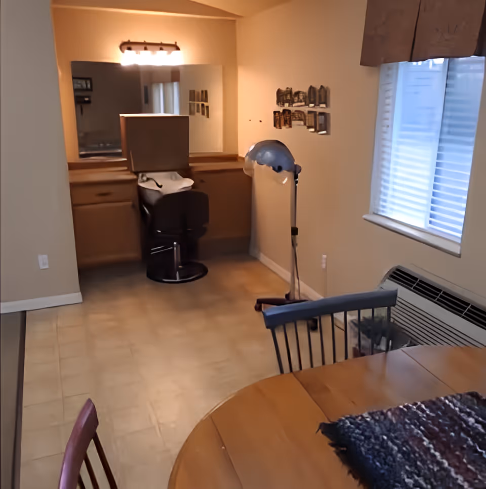 Interior room with a wooden dining table and chairs in the foreground, a window with blinds on the right wall, and a vanity area with a large mirror, chair, and hair dryer in the background.