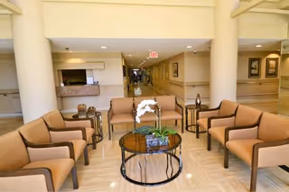 A spacious lobby seating area with upholstered armchairs arranged around a glass coffee table and a hallway leading further into the facility.
