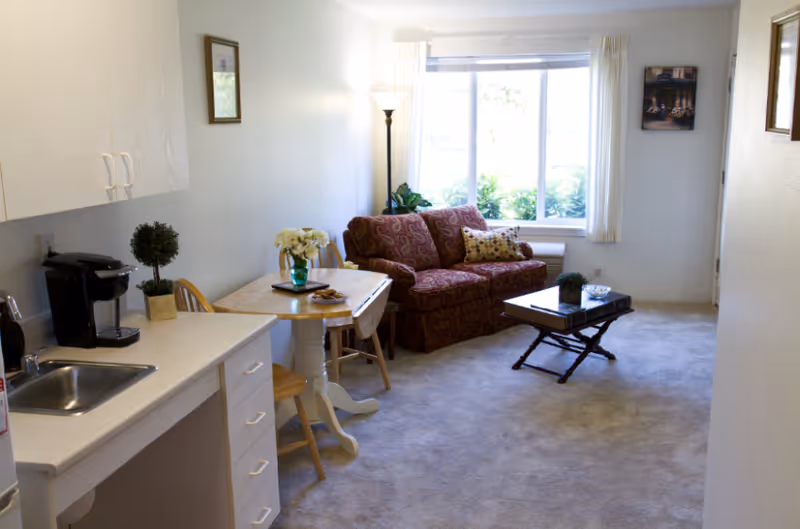A cozy living area in a retirement facility featuring a small kitchen counter with a sink and coffee maker on the left, a wooden dining table with chairs and a vase of flowers, a patterned red loveseat with decorative pillows, a floor lamp, a coffee table with a small plant, and a large window with white curtains letting in natural light.