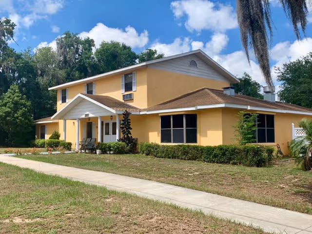 A two-story yellow building with a brown roof, white trim, and a covered entrance with white columns. The building is surrounded by green bushes and trees, with a concrete sidewalk leading up to the entrance. The sky is blue with some clouds.