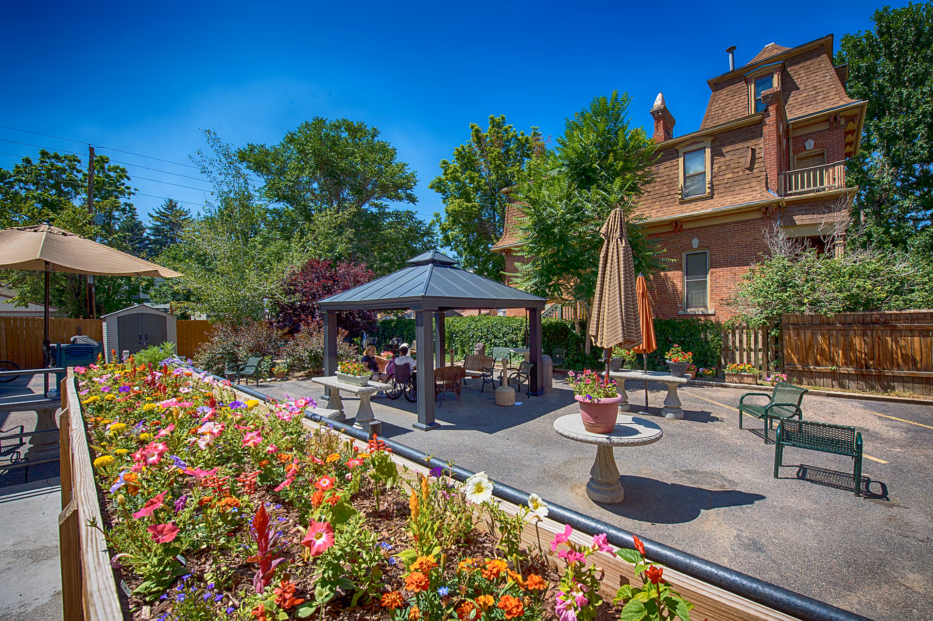 Outdoor garden area at Denver North Care Center with colorful flowers in raised beds, a gazebo with seating underneath, benches, tables with umbrellas, and a large brick building in the background under a clear blue sky.