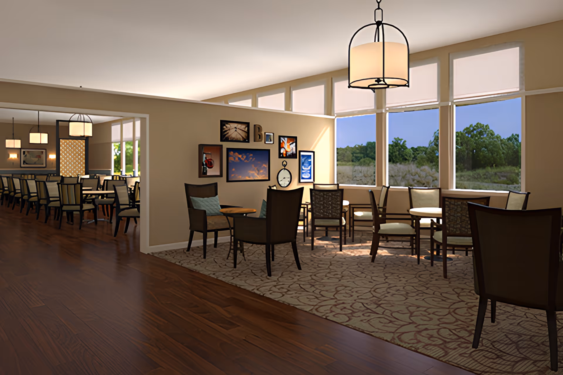 A bright and spacious common area in a senior living facility with large windows showing a view of trees outside. The room features several seating arrangements with chairs and small tables on a patterned carpet, and a wooden floor leading to a dining area with multiple tables and chairs. The walls are decorated with framed pictures and a clock, and a hanging light fixture is visible.