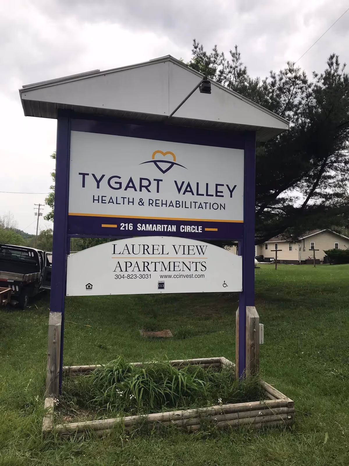 Outdoor sign for Tygart Valley Health & Rehabilitation located at 216 Samaritan Circle, with an additional sign below for Laurel View Apartments including a phone number and website. The sign is purple and white, set on a grassy area with trees and a building in the background.