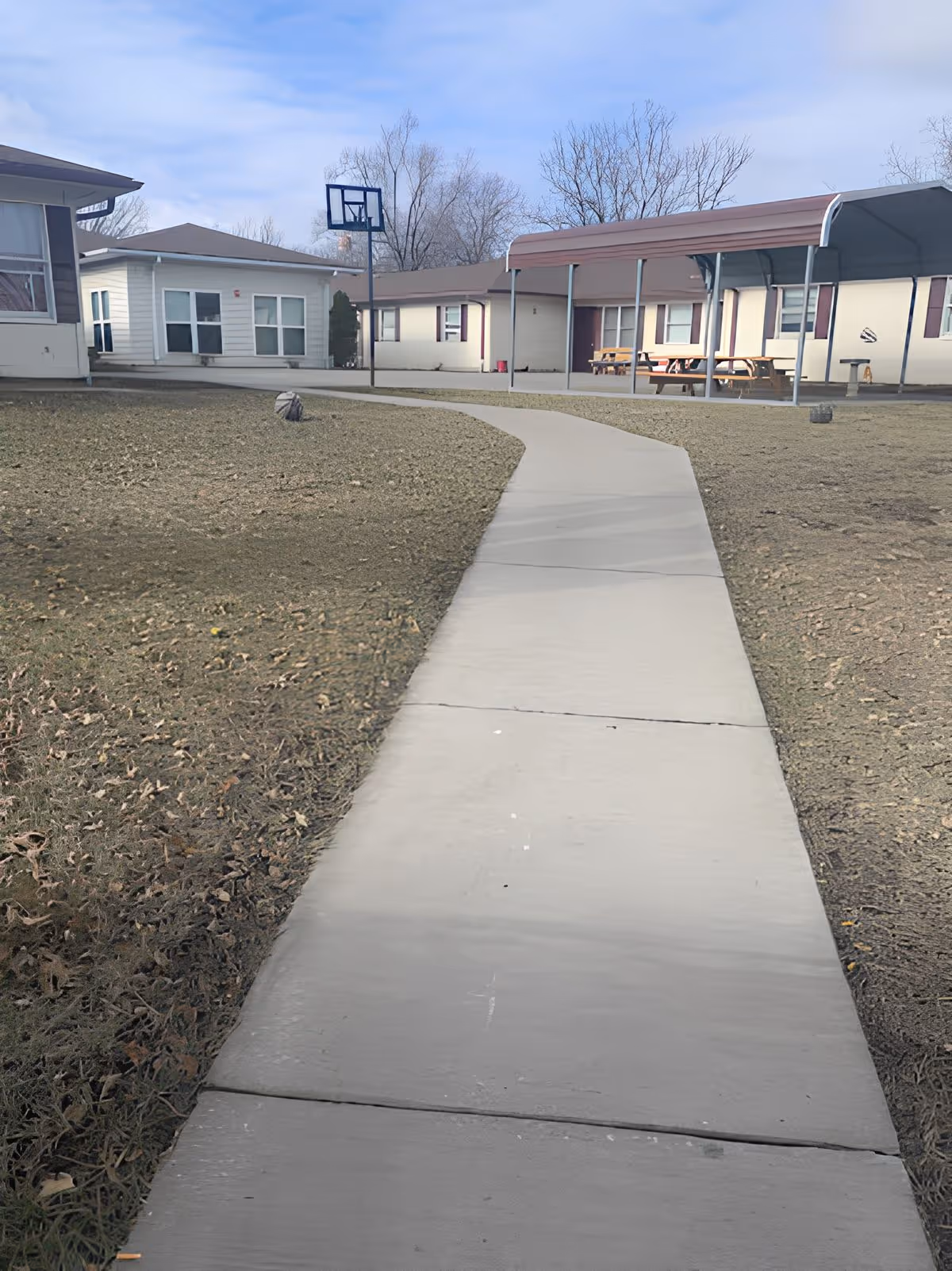 Concrete walkway leading through a courtyard toward single-story assisted living buildings with a covered picnic area and a basketball hoop.