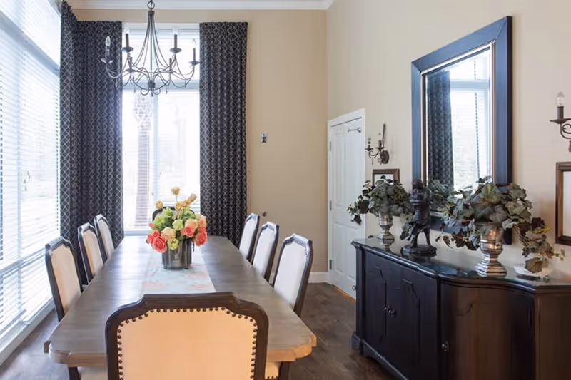 Bright formal dining room with a long wooden table surrounded by upholstered chairs, a chandelier, large windows with drapes, and a sideboard topped with a mirror and plants.