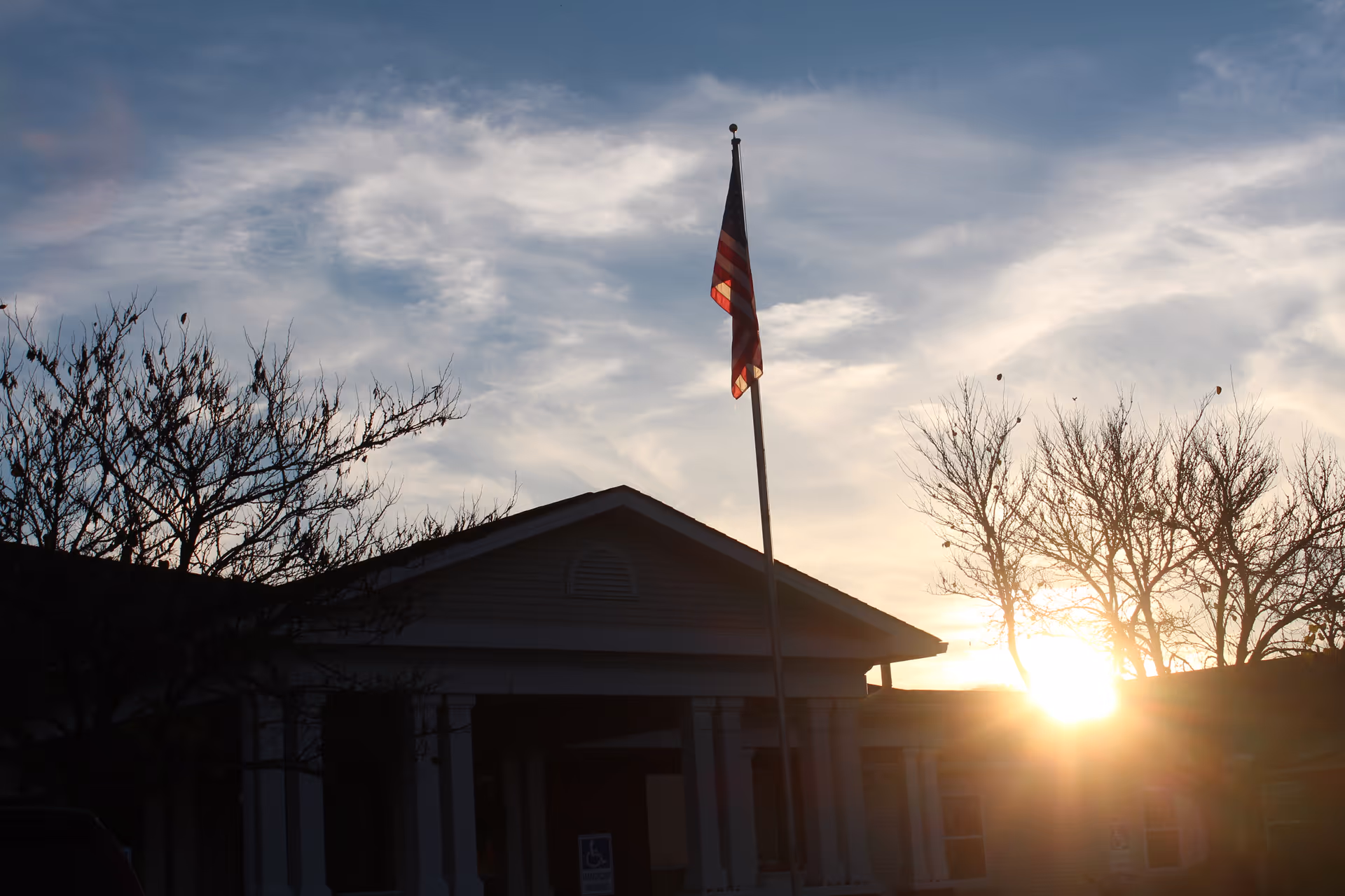 Front of a building silhouetted against a setting sun with an American flag on a pole and bare trees.