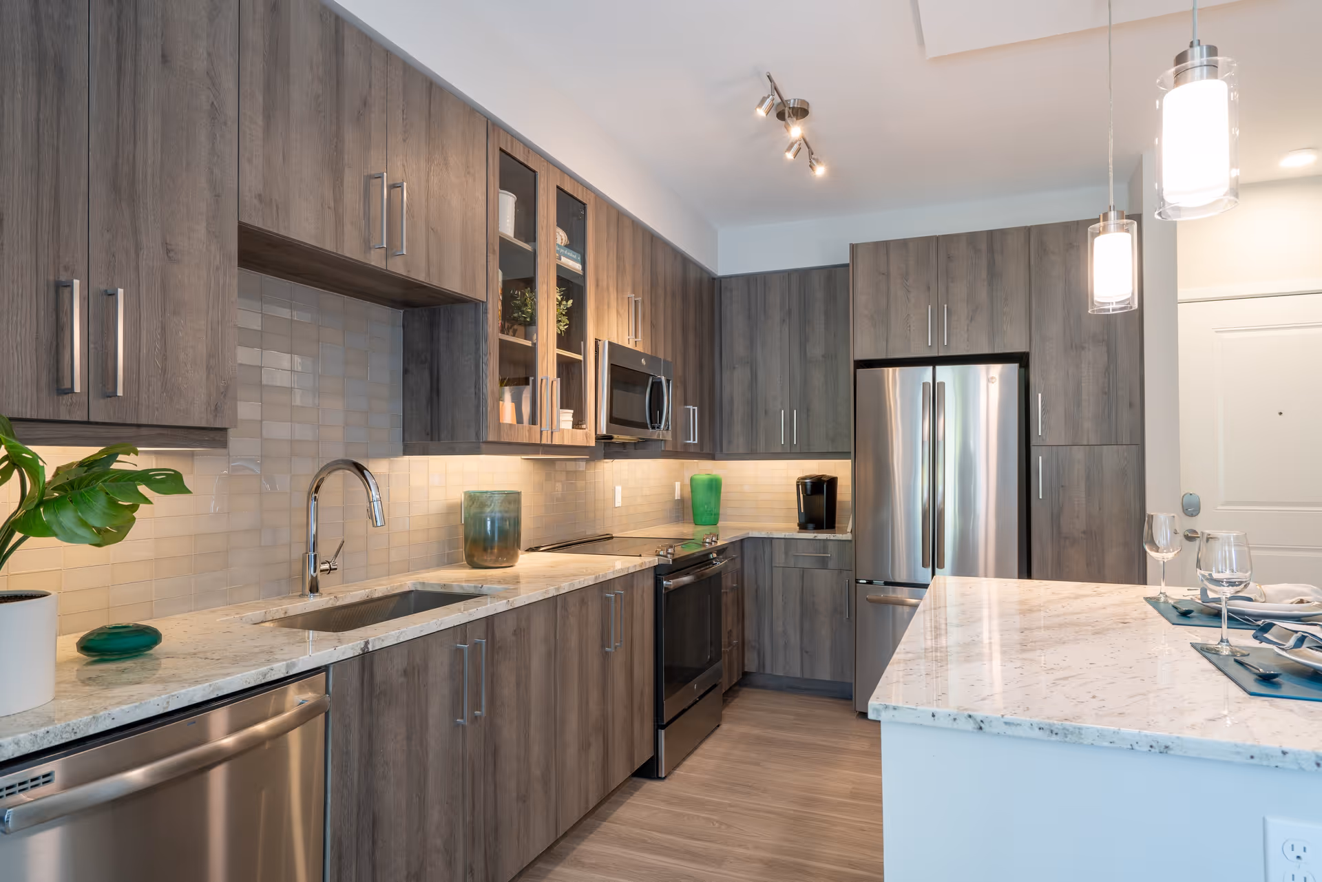 Modern kitchen with wood-finish cabinets, stainless steel appliances including a refrigerator, oven, microwave, and dishwasher. The kitchen features a light-colored granite countertop with a sink, under-cabinet lighting, and two pendant lights hanging over a breakfast bar set with plates, napkins, and wine glasses.