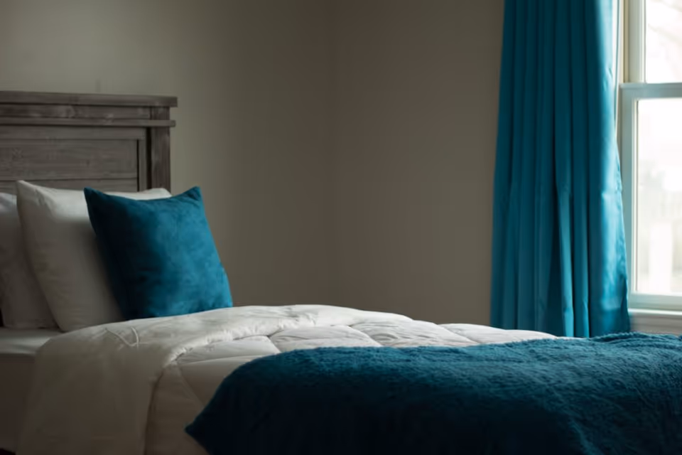 A neatly made bed with white sheets, a teal throw blanket, and a teal pillow against a wooden headboard. There is a window with teal curtains letting in natural light.