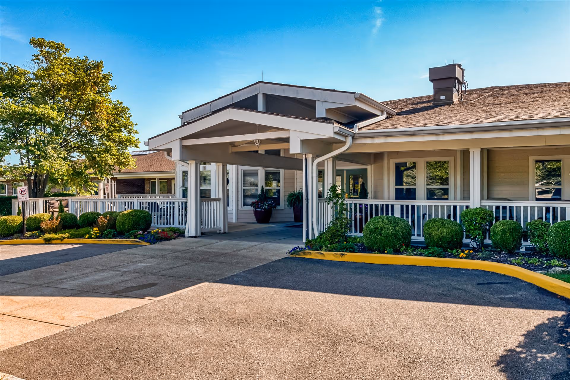 Front entrance of a single-story senior living facility with a covered porte-cochere, white railing, landscaped shrubs and a driveway.