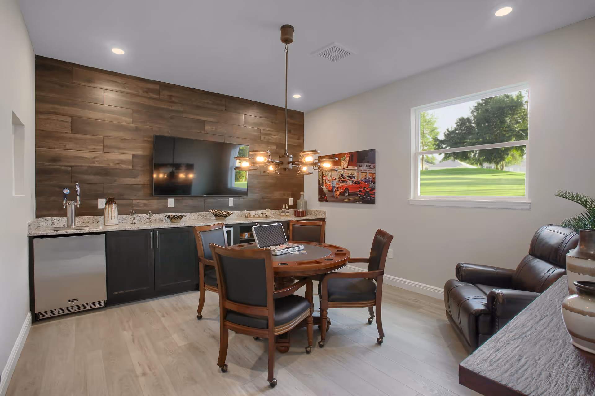 Small living/entertainment room with a round wooden game table and chairs, leather recliner, wet bar with TV on a wood-paneled wall, and a window showing a green lawn.