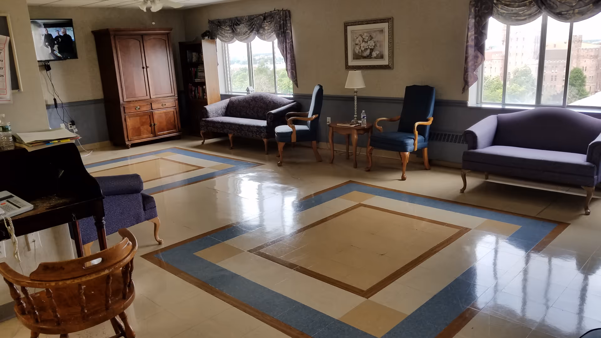 Sunlit common lounge with upholstered sofas and chairs arranged around a decorative tiled floor and large windows.