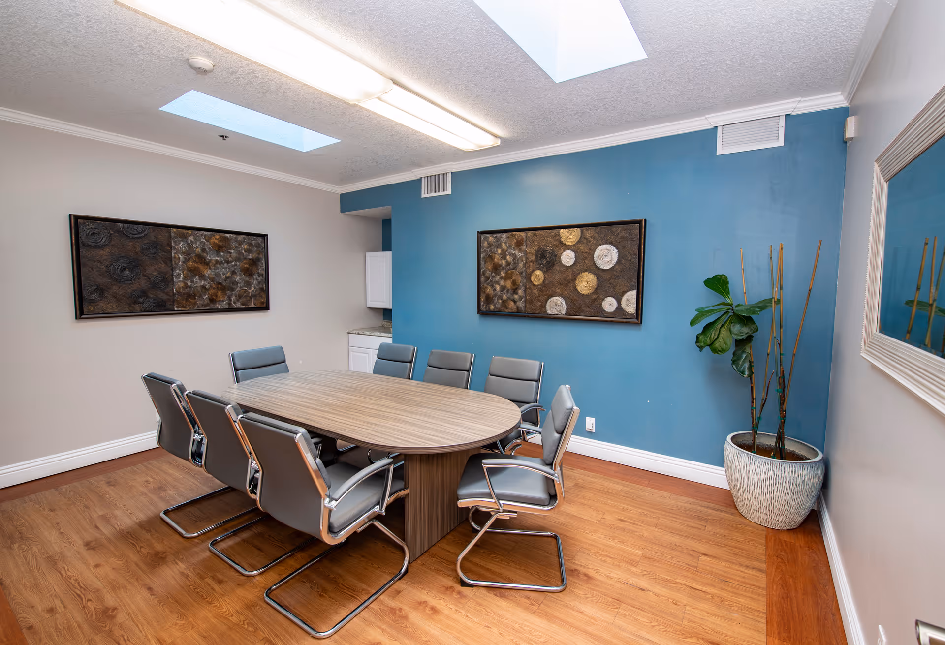 A modern conference room with a wooden oval table surrounded by eight gray cushioned chairs with metal frames. The room has wooden flooring, a blue accent wall with a framed abstract art piece, a large potted plant in the corner, and two skylights on the ceiling providing natural light. There is also a large mirror on the adjacent wall and a small white cabinet in the corner.
