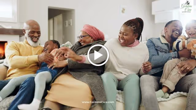 A joyful multi-generational family sitting closely together on a couch in a living room, laughing and playing with each other. The group includes older adults, a middle-aged woman, and two young children. The setting is cozy and bright.
