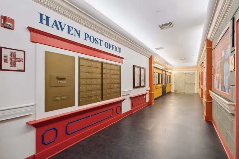 Interior hallway of The Haven and The Laurels in Stone Oak featuring a wall-mounted post office area with multiple mailboxes labeled 'HAVEN POST OFFICE'. The hallway has decorative molding, framed pictures, and a clean, well-lit environment with a door at the far end.