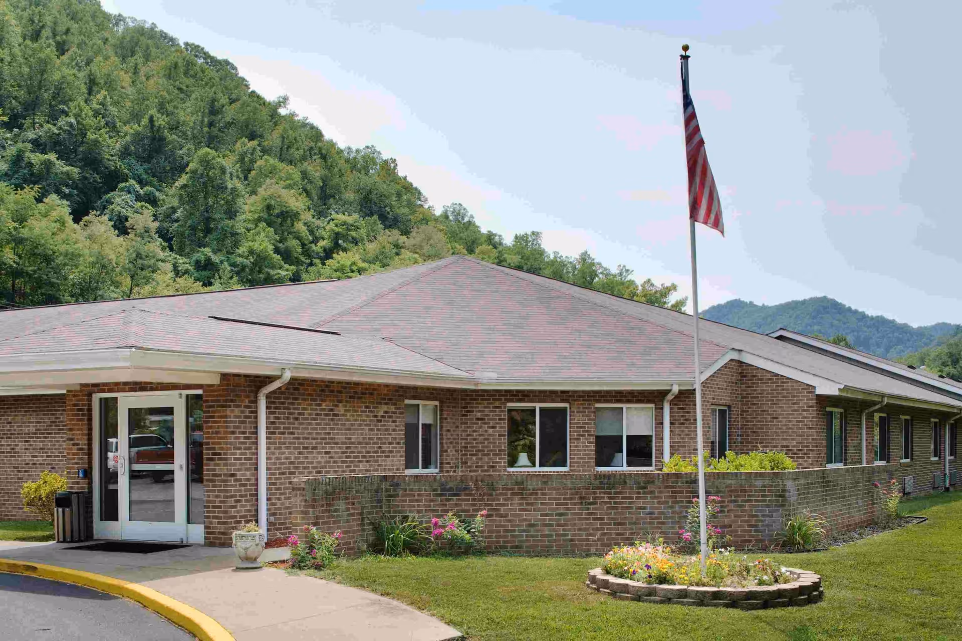 Exterior view of a single-story brick building with a sloped roof, surrounded by green grass and trees. An American flag is flying on a flagpole in front of the building, which has several windows and a glass entrance door. Hills covered with trees are visible in the background.