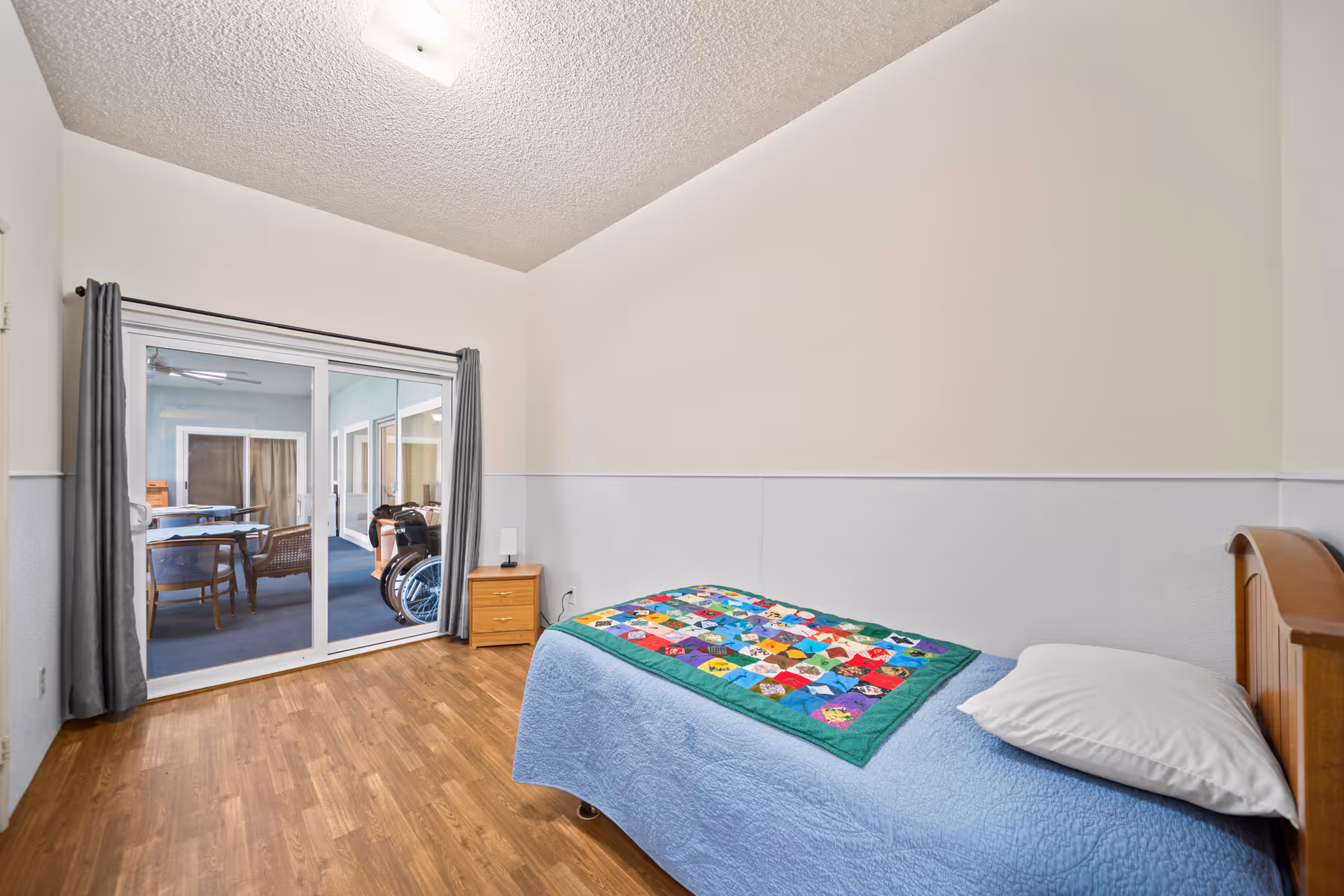 A simple bedroom with a single bed covered with a blue quilt and a colorful patchwork blanket. There is a wooden nightstand with a small lamp next to the bed. The room has wood flooring and white walls with a textured ceiling. Sliding glass doors with gray curtains lead to an enclosed patio area with chairs and a wheelchair visible outside.