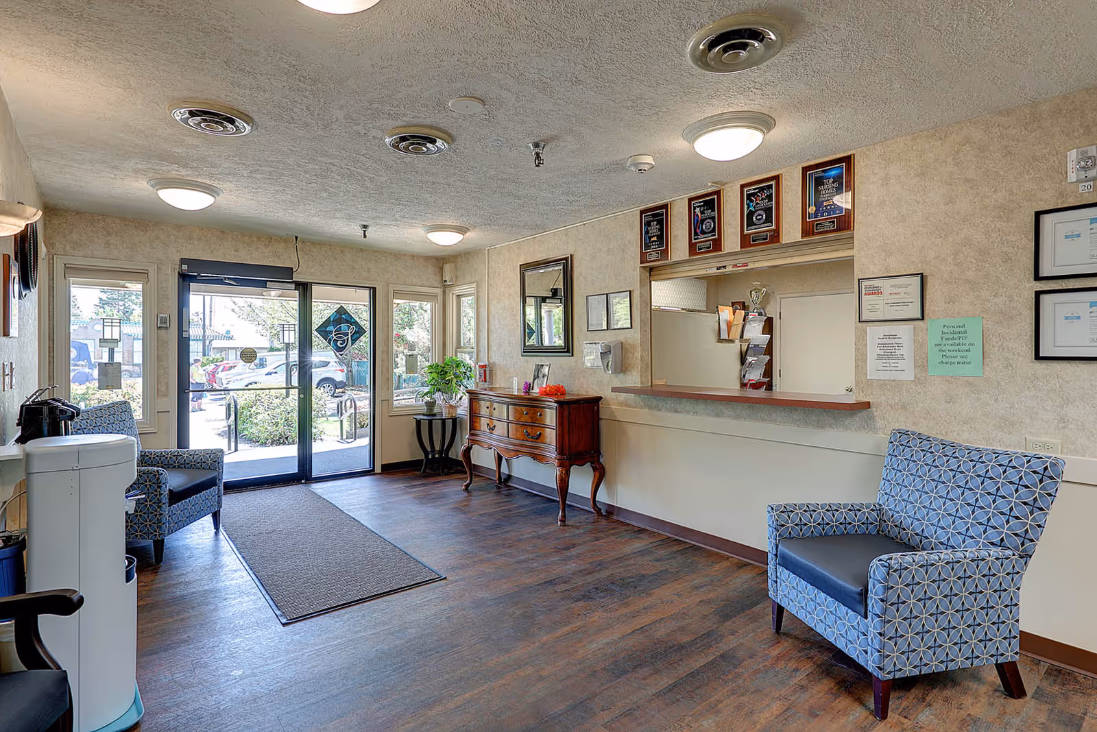 Reception area of Gateway Care & Retirement Center with a wooden floor, two patterned armchairs, a wooden sideboard with a plant and decorations, a reception window with plaques above it, and glass doors leading outside.