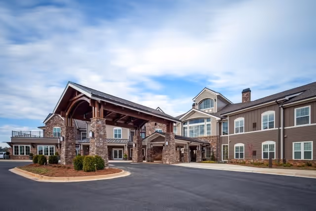 Exterior view of a senior living facility building with a large covered entrance supported by stone pillars, multiple windows, and a paved driveway under a partly cloudy sky.