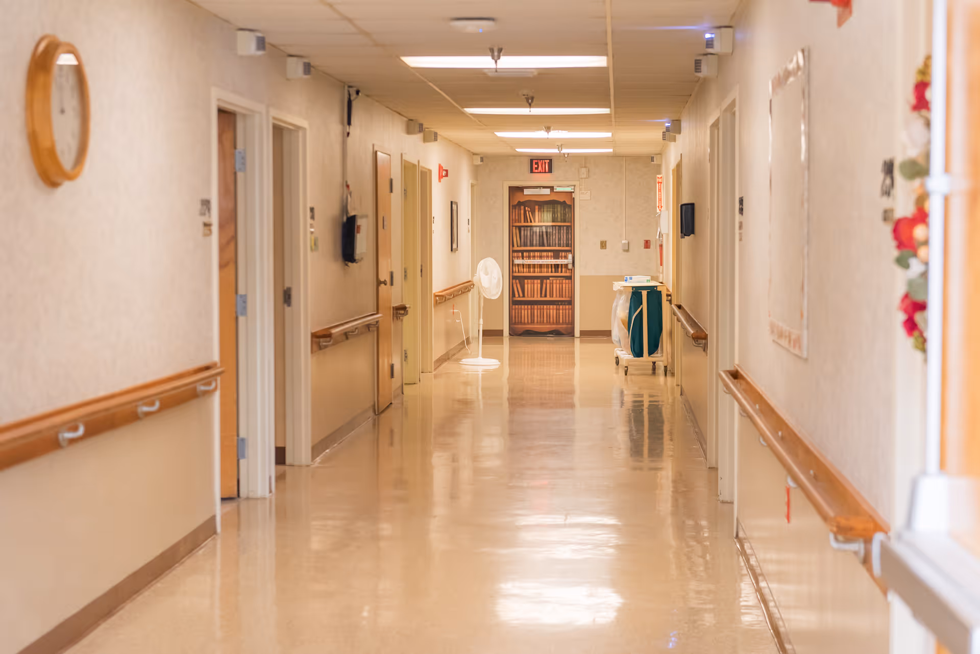 Well-lit empty hallway in a senior living facility with handrails along the walls, doors on either side, and a fan and cart at the far end.