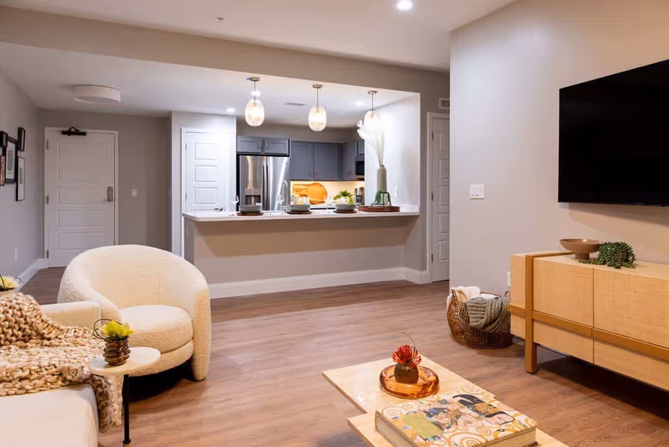 A modern and cozy living room area with a white armchair, a beige sofa with a chunky knit blanket, a small side table with a plant, and a wooden coffee table with a decorative tray and book. The room has wood flooring and neutral-colored walls. In the background, there is an open kitchen with gray cabinets, stainless steel refrigerator, and three pendant lights hanging over a breakfast bar with place settings. A flat-screen TV is mounted on the wall above a wooden console with decorative items.