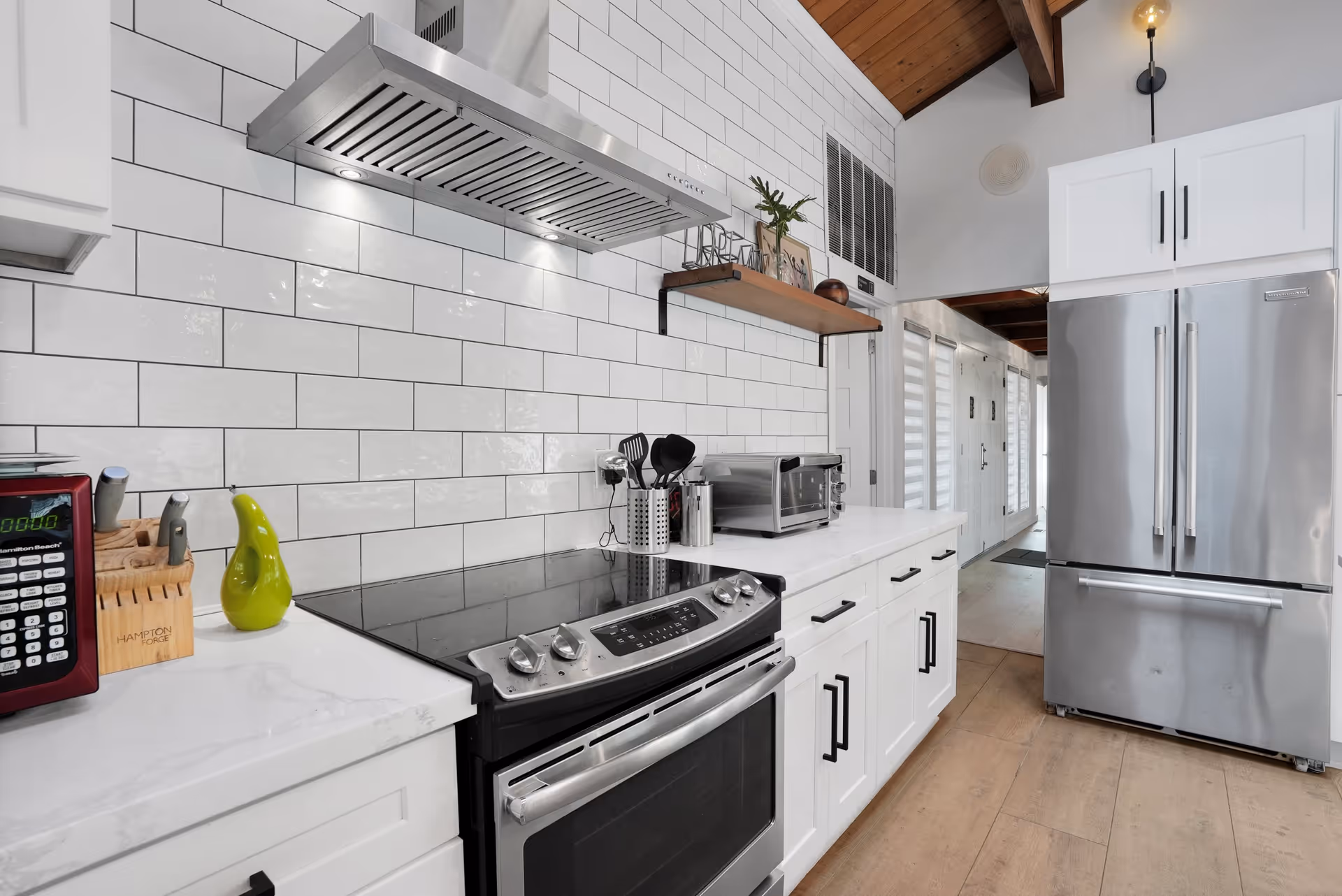 Modern kitchen with white cabinets, a stainless steel stove and oven, a stainless steel refrigerator, a microwave, a knife block, and a small green pear-shaped decoration on the white countertop. The backsplash is white subway tile, and there is a wooden shelf with decorative items above the counter. The ceiling has wooden beams and a hanging light fixture.