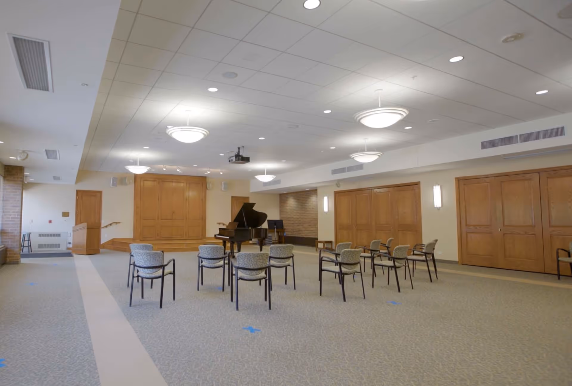 A spacious room with a small stage area featuring a grand piano and a music stand. Several chairs are arranged facing the stage. The room has a carpeted floor, wooden panel doors, and ceiling lights providing bright illumination.