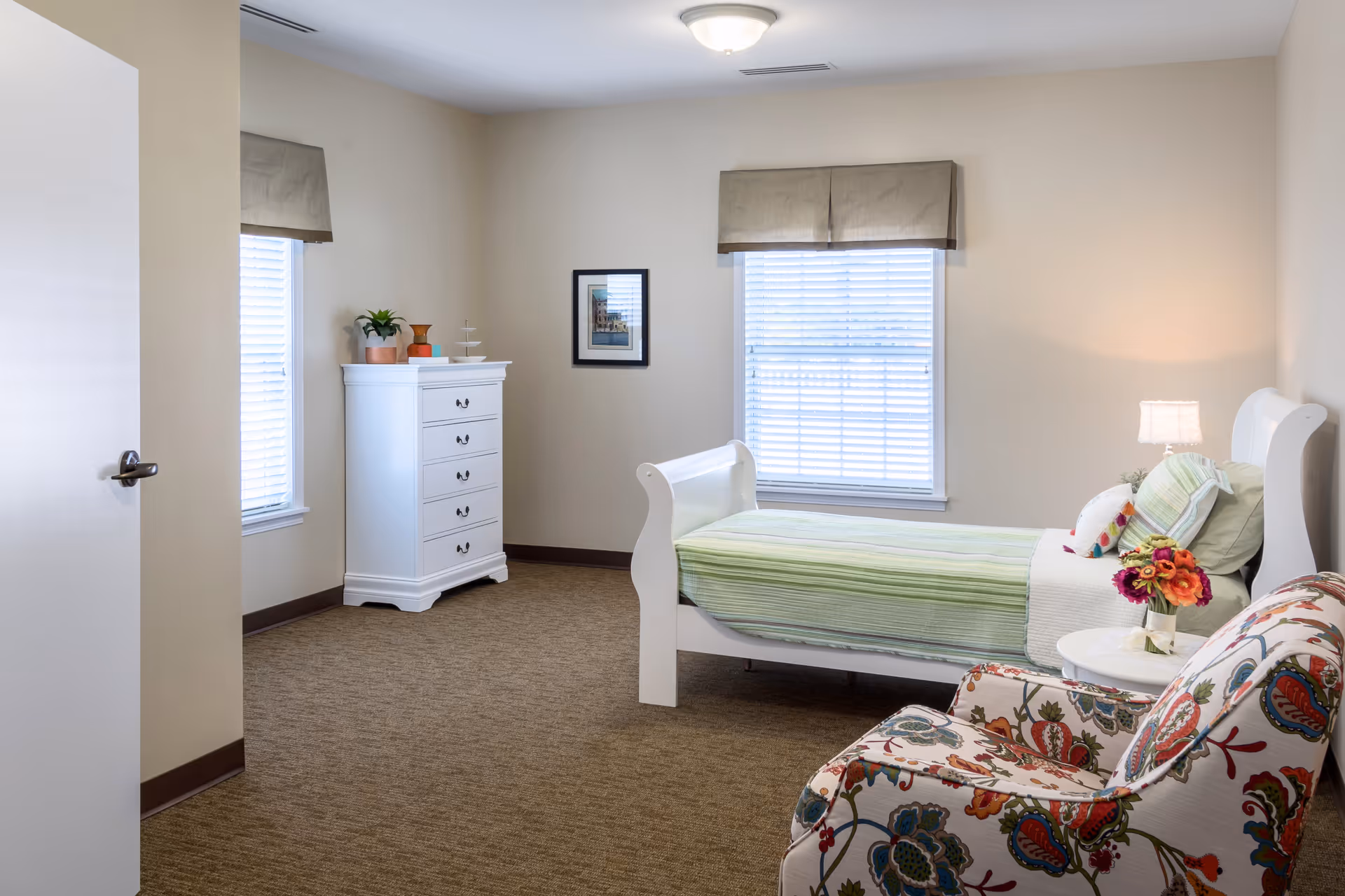 A bright and tidy bedroom with a single white bed covered in green striped bedding, a floral patterned armchair, a small round side table with a vase of flowers, a white chest of drawers, two windows with blinds and beige valances, and a framed picture on the wall.