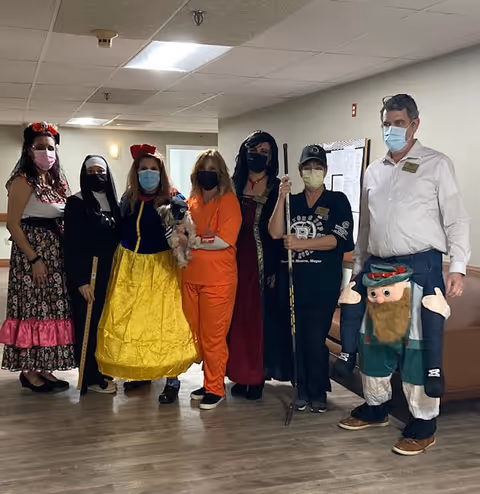 A group of seven people standing indoors in a hallway. They are dressed in various costumes including a floral dress, nun outfit, Snow White, orange scrubs holding a small dog, a medieval-style dress, casual black outfit with a cap holding a broom, and a man wearing a costume that makes it look like a small person is carrying him. All individuals are wearing face masks.