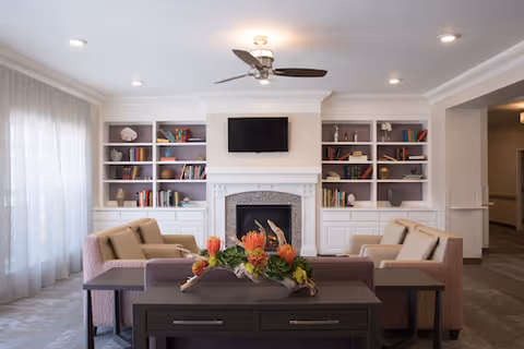 A cozy living room area with two beige armchairs and a sofa arranged around a dark wooden coffee table with a floral centerpiece. Behind the seating is a white fireplace with a mounted flat-screen TV above it, flanked by built-in bookshelves filled with books and decorative items. The room is softly lit with recessed ceiling lights and a ceiling fan with a light fixture. Sheer curtains cover the large window on the left side.