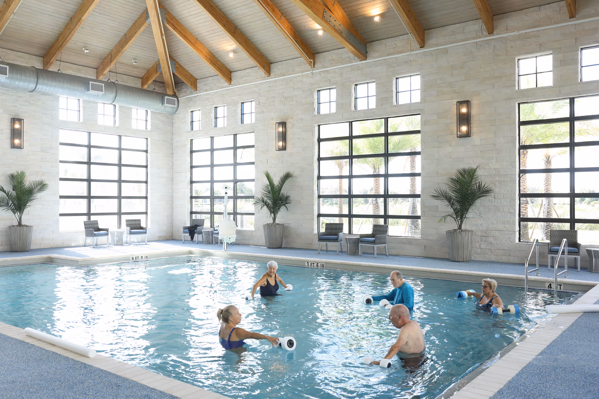 Indoor swimming pool with five elderly people participating in water exercise using foam dumbbells. The room has large windows with views of palm trees outside, high ceilings with exposed wooden beams, and potted plants along the walls. Several chairs and small tables are placed near the windows.