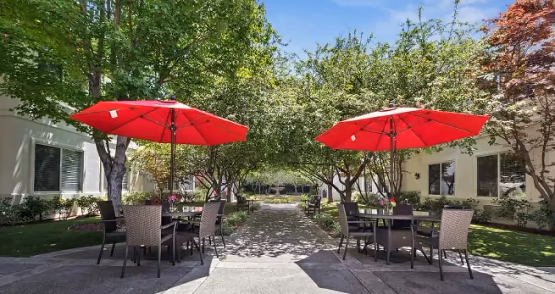 Outdoor courtyard area with two tables each shaded by a large red umbrella, surrounded by chairs. The area is bordered by trees and shrubs with a clear blue sky overhead.