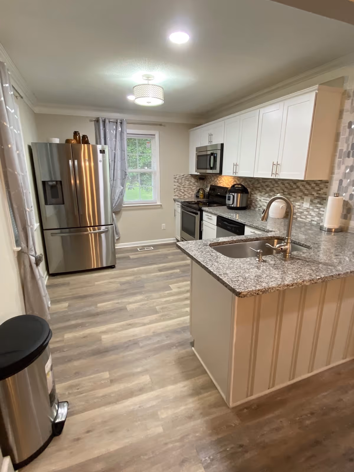 A modern kitchen with light wood flooring, white cabinets, and granite countertops. The kitchen features a stainless steel refrigerator, microwave, stove, dishwasher, and a sink with a curved faucet on the island. There is a window with gray curtains and a trash can near the entrance.
