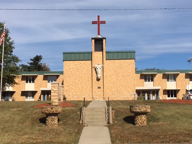 Front exterior of a brick senior care building with a red cross and a statue above the entrance and steps leading up the lawn.