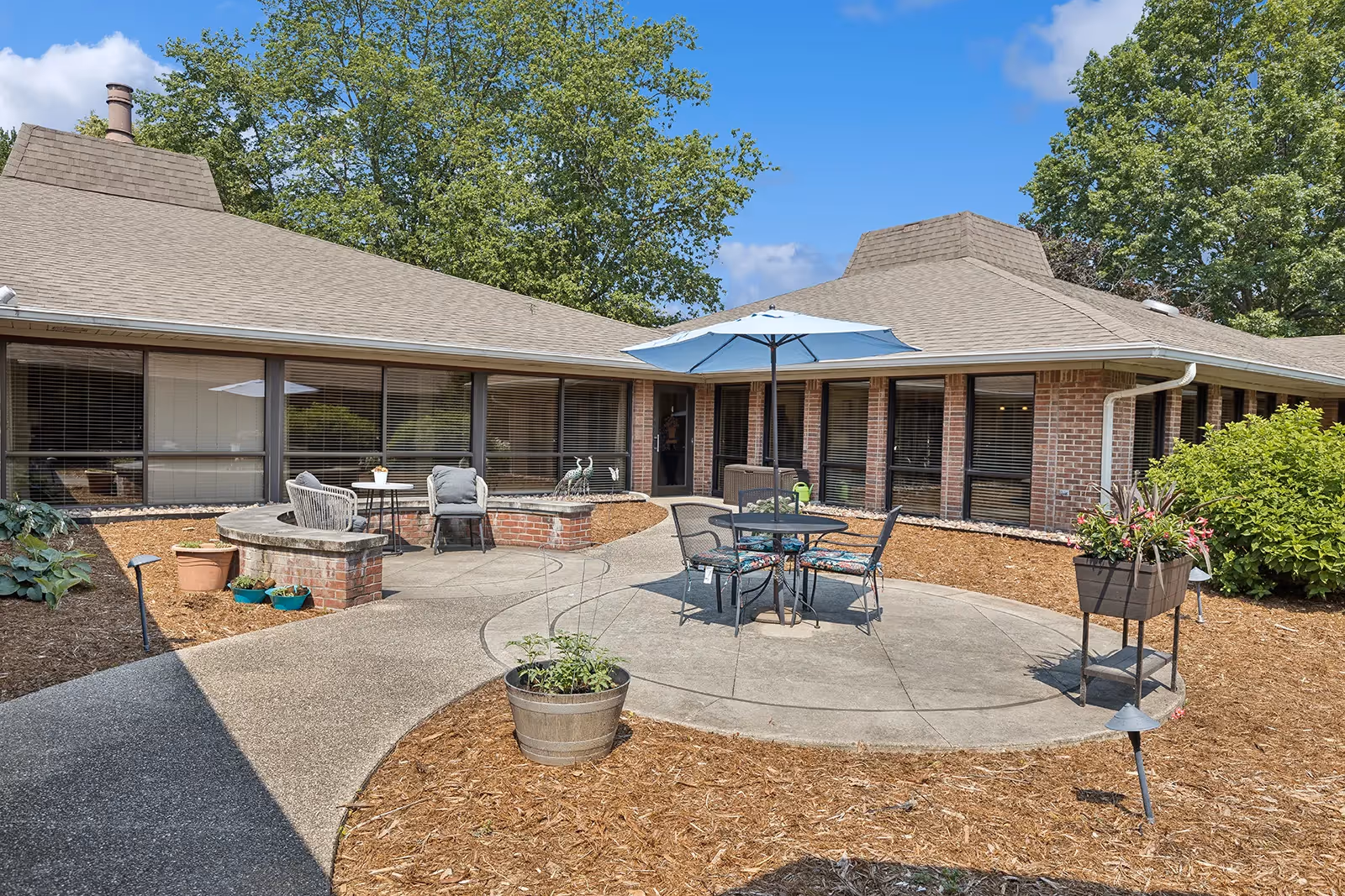 Outdoor patio area of a senior living facility with a round table and four chairs under a blue umbrella. The patio is surrounded by a brick building with large windows and landscaping including potted plants and mulch beds. Trees and a blue sky with some clouds are visible in the background.