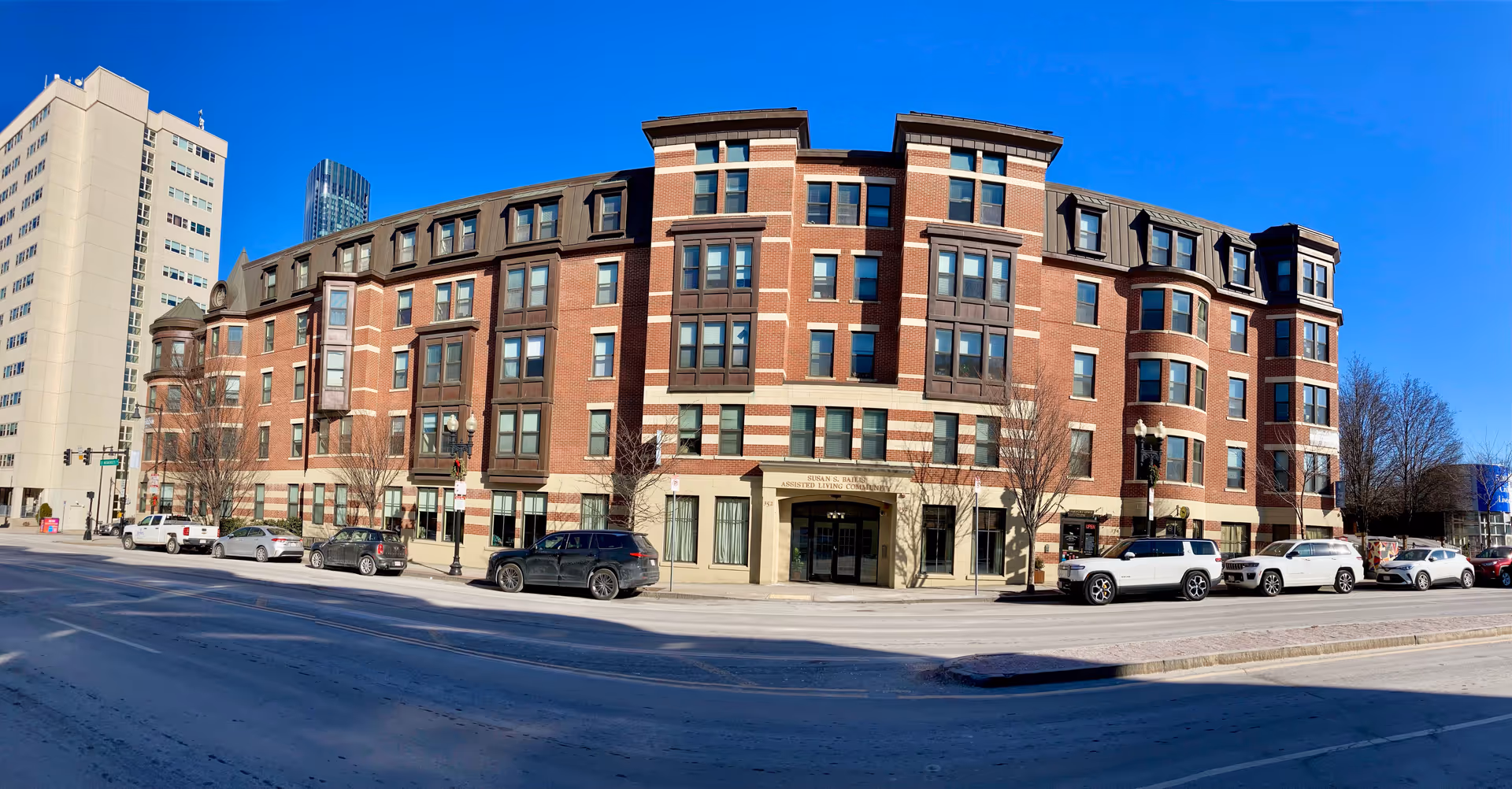 Front exterior of the Susan S. Bailis Assisted Living brick building on a sunny day with cars parked along the street.