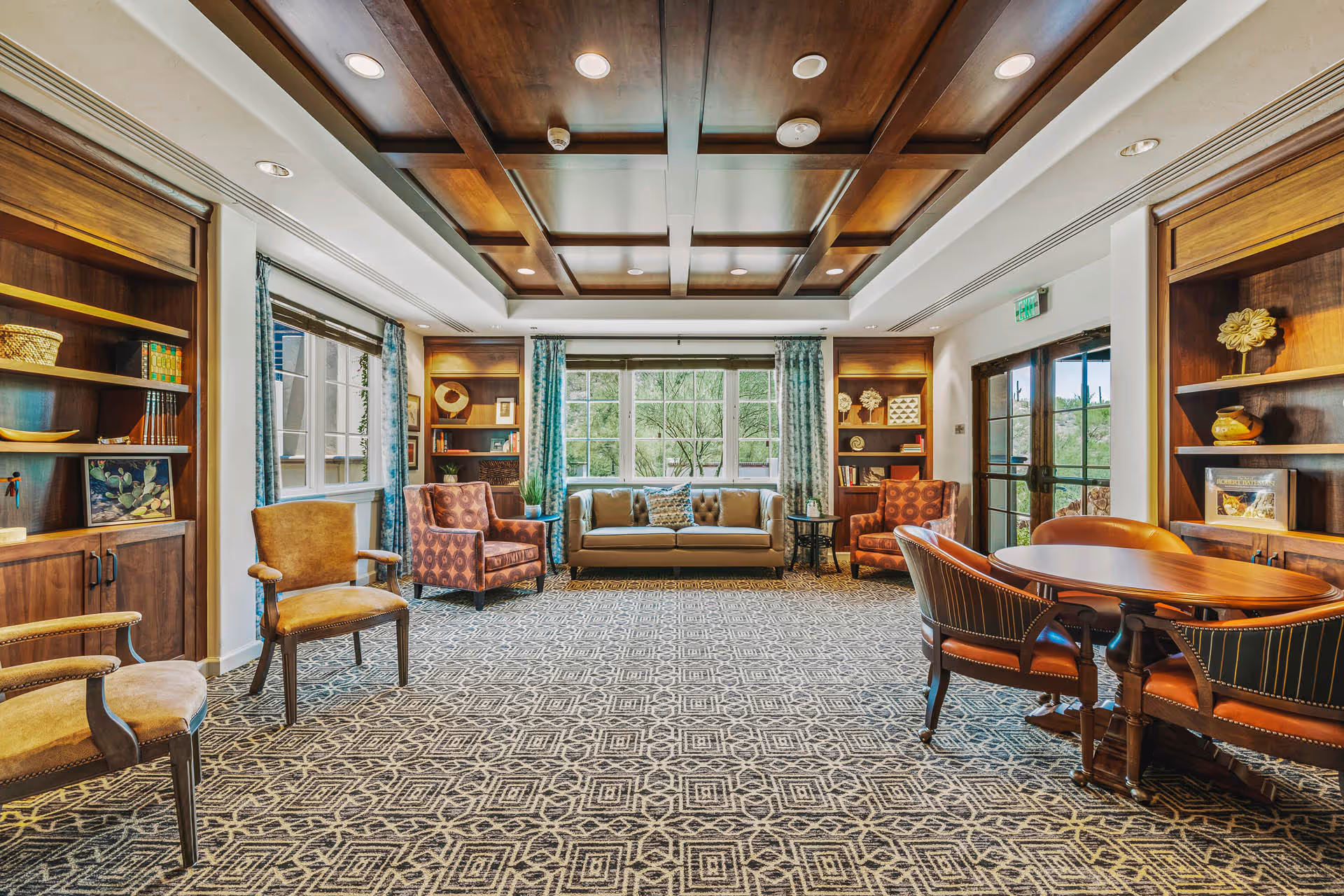 A spacious and well-lit living room area with a patterned carpet, wooden coffered ceiling, and large windows with blue curtains. The room features a beige sofa, several upholstered armchairs, wooden bookshelves with decorative items, and a round wooden table with chairs near a glass door leading outside.