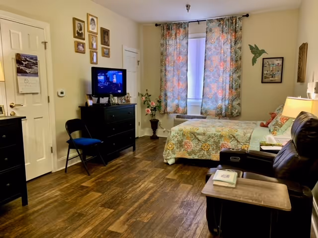 A cozy bedroom with a bed covered in a floral quilt, a window with floral curtains, a black dresser with a TV on top, a black folding chair, a leather recliner with a side table, and framed pictures on the walls.