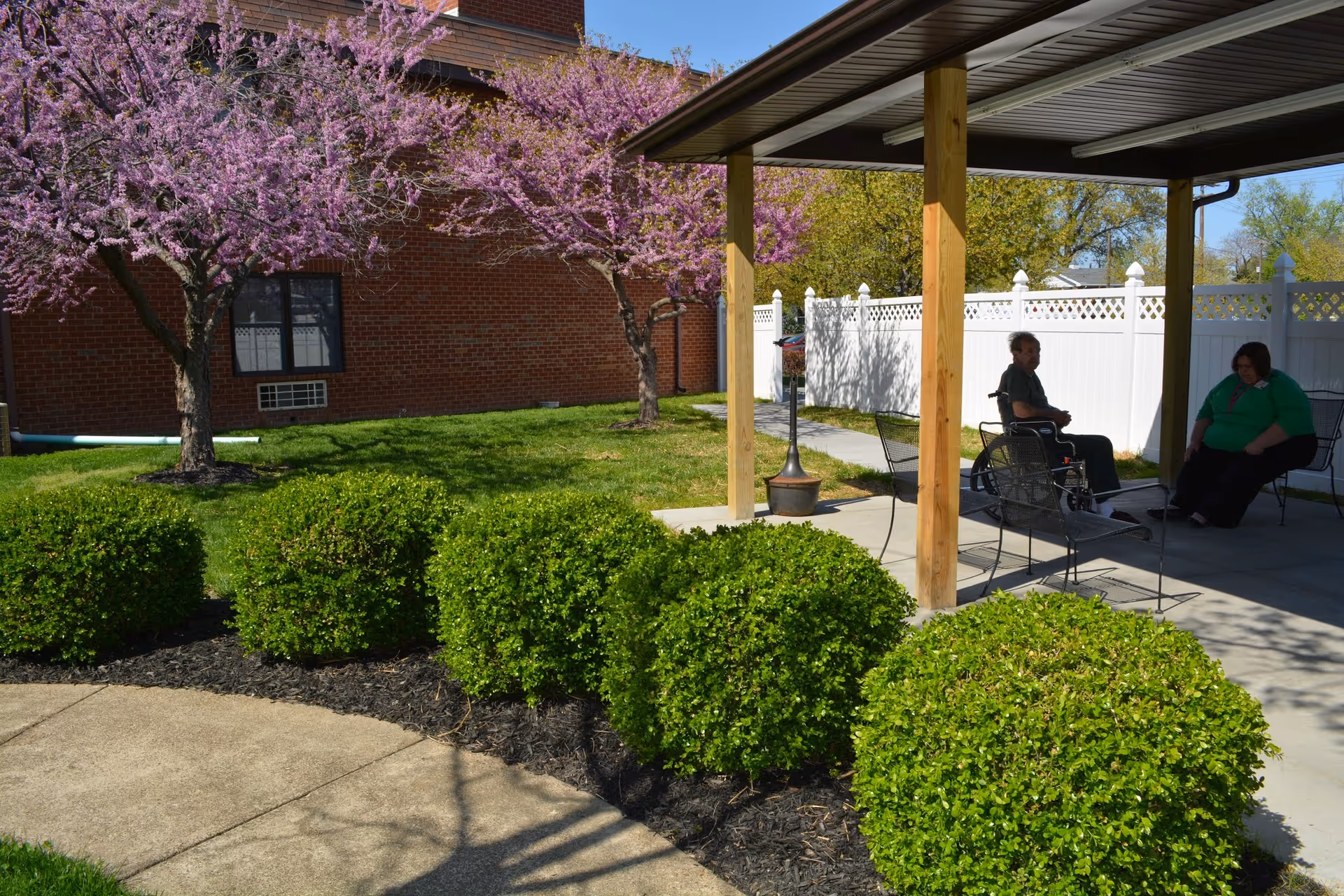 Outdoor seating area with a wooden pergola, two people sitting on metal chairs, green bushes, blooming purple trees, and a brick building in the background.