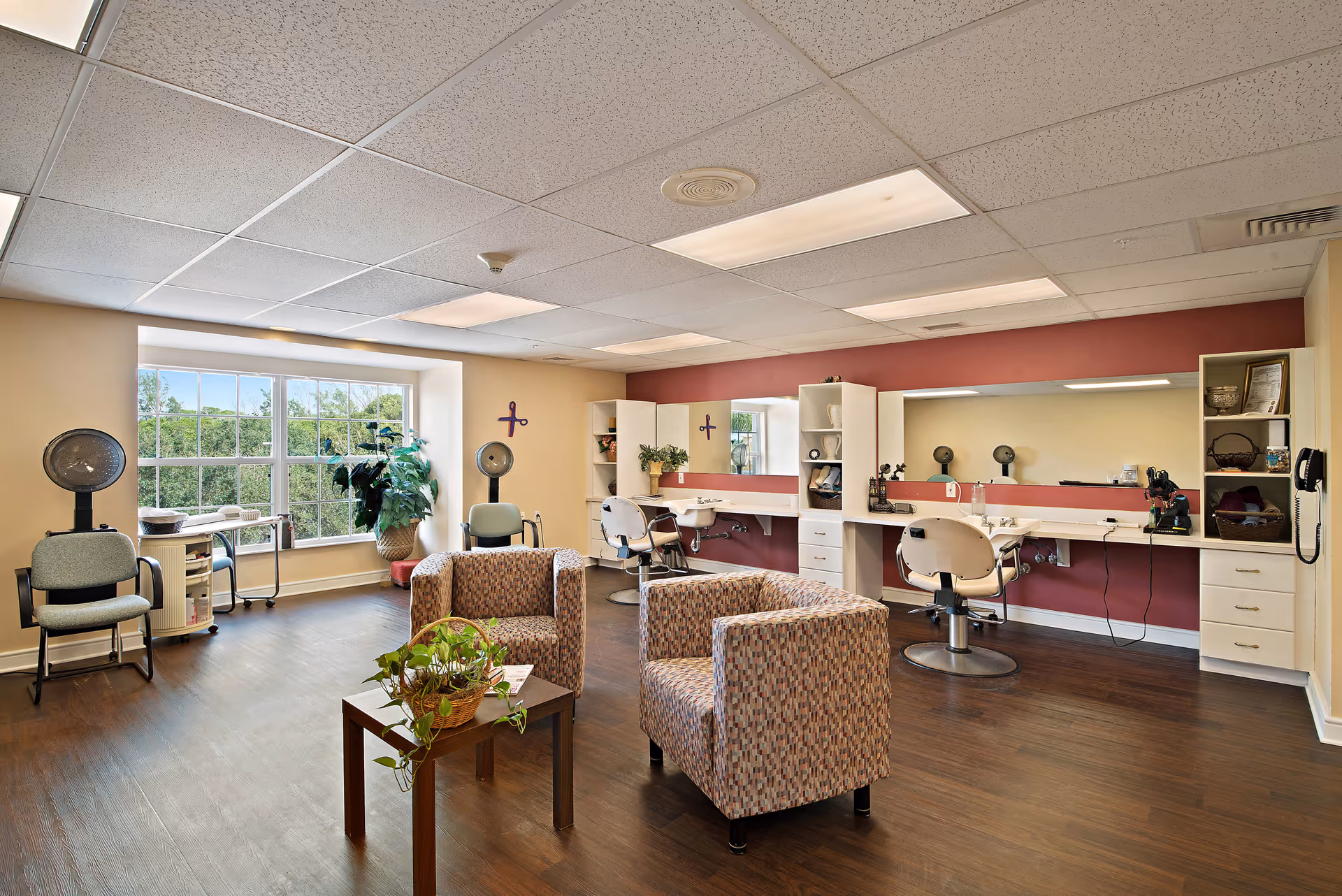 A bright and spacious hair salon area in a senior living facility with two patterned armchairs around a small wooden table with a plant. The room has large windows letting in natural light, salon chairs in front of mirrors, hair dryers, and shelves with decorative items and supplies. The walls are painted beige and maroon, and the floor is dark wood.