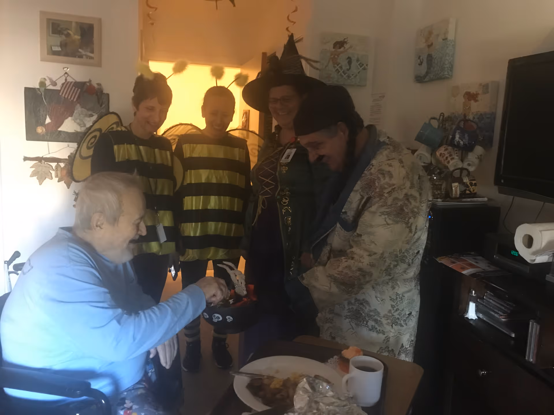 An elderly man in a wheelchair reaching into a Halloween-themed container held by a person dressed in a patterned robe and hat. Behind them, two people are dressed in bee costumes and another person is dressed as a witch. The setting appears to be a cozy room with a table holding a plate of food and a cup, and various decorations on the walls.