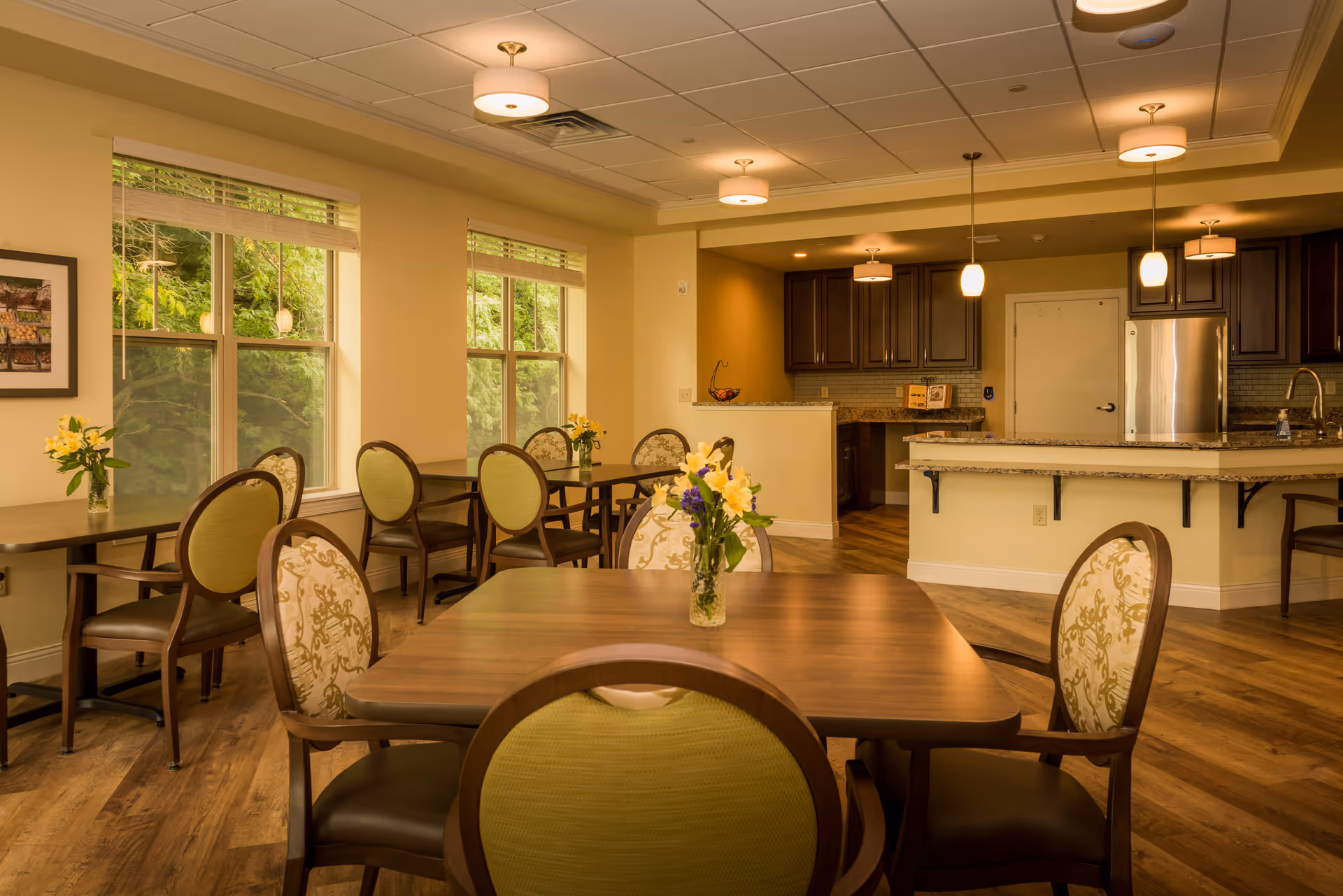 Spacious dining area with wooden tables and upholstered chairs, floral centerpieces, large windows, and an open kitchen in the background.