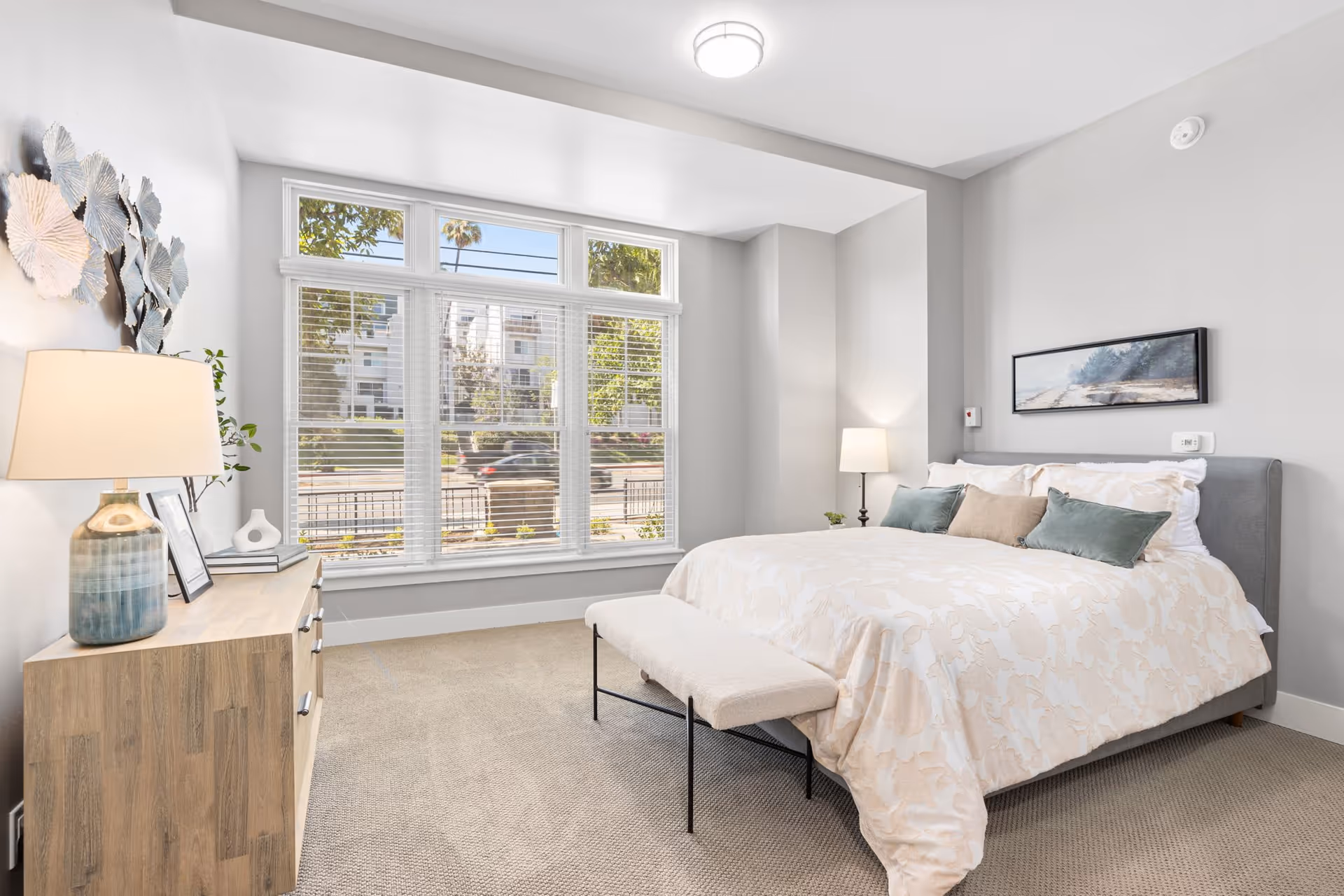 A bright and spacious bedroom with a large window letting in natural light. The room features a neatly made bed with white and beige bedding and several pillows, a cushioned bench at the foot of the bed, a wooden dresser with a lamp and decorative items, and a wall-mounted landscape painting. The walls are painted light gray and the floor is carpeted.
