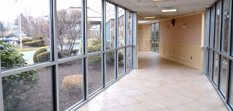 A bright indoor hallway with large floor-to-ceiling windows on both sides, showing landscaped bushes and trees outside. The hallway has beige tiled flooring, cream-colored wainscoting on the walls, and a light pink upper wall section with two wall sconces. The ceiling has recessed lighting panels.